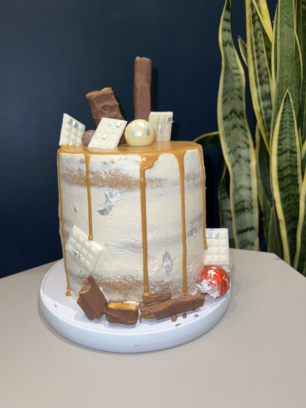 A tall semi-naked celebration cake with salted caramel drip, topped with chocolate bars, wafers and truffles
